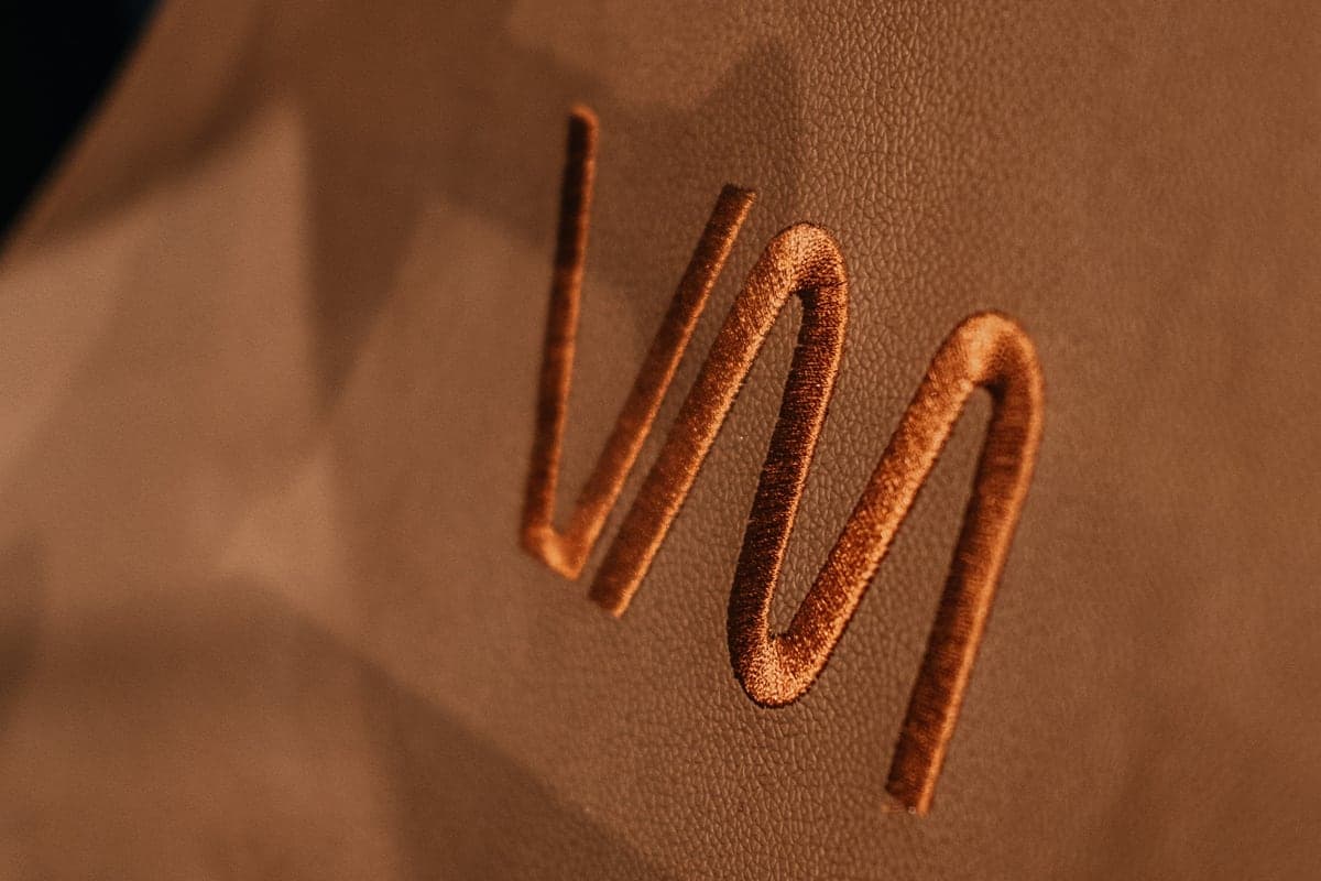 Detalhe do símbolo “M” da marca Via Move em relevo, cor cobre.